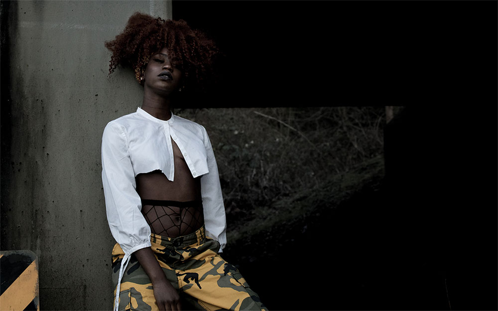 Black woman with curly, maroon hair 
            standing with her back to a stone wall. Her legs are slightly bent and she is facing the camera.
            Woman is wearing yellow and black camouflage pants and a white cut-off button up shirt.