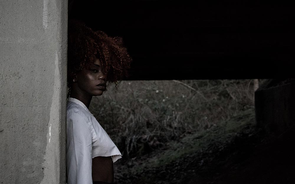 Black woman with curly, maroon hair standing 
            with her back to a stone wall. Her body is faced to the right, while her 
            head is turned towards the camera.